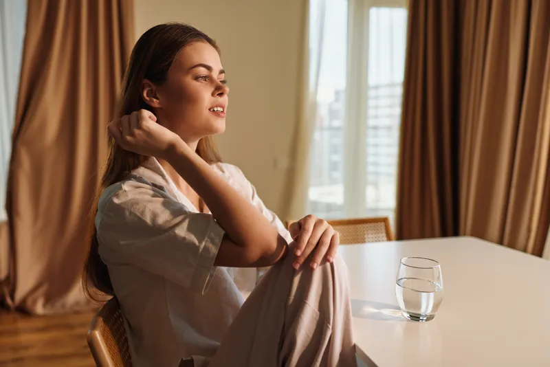 Woman portrait at home relaxing on a chair by a table with a glass of water, soft sunlight through curtains, casual outfit and contemplative expression in warm living space.