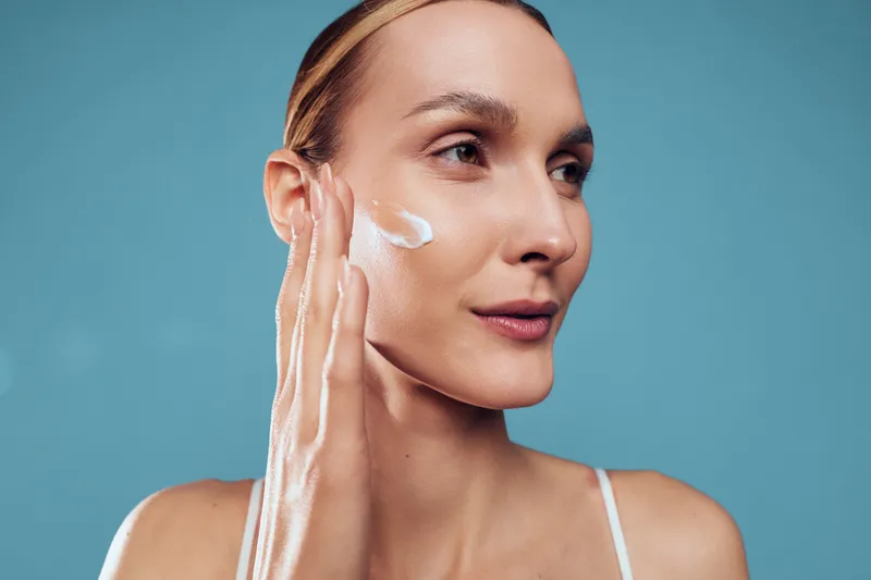 Woman applies skincare cream on face, beauty treatment for hydration, spa selfcare with cosmetic texture, serene expression, soft closeup portrait, blue studio background, clean skin