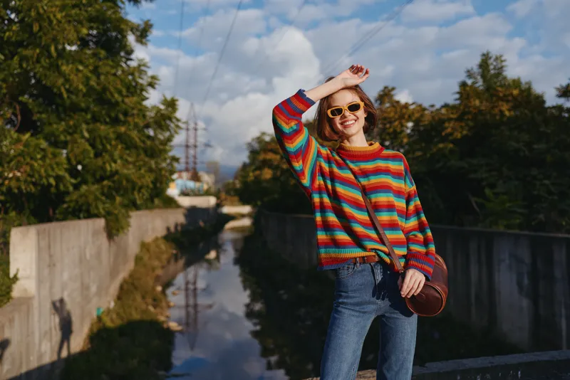 Young woman in her 20s wearing a colorful striped sweater and sunglasses, smiling and leaning on a railing by an urban canal, casual jeans and crossbody bag for lifestyle travel portrait.