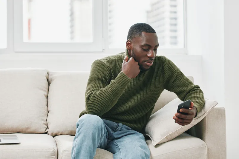 Thoughtful young Black man in casual green sweater checking smartphone while sitting on cozy sofa in modern living room, reflecting on life choices and goals