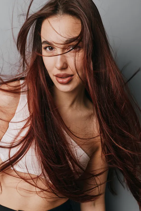 Young woman with long red hair and natural makeup posing in white top. Closeup portrait with hair flowing and soft expression against neutral background in studio lighting.