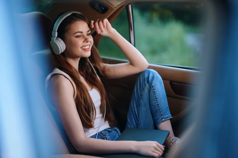 Young woman enjoying music in a car, wearing headphones, sunny day, relaxed and happy, casual outfit with jeans, concept of freedom and leisure.