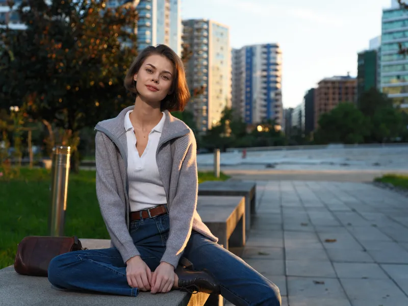 A relaxed young woman sits on a bench in a modern city park, dressed casually in jeans and a light cardigan. She gazes calmly toward the camera amid urban scenery.