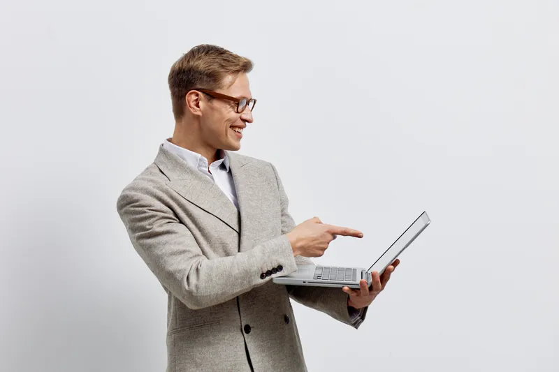 Smiling man in smart casual attire holding a laptop and pointing at the screen against plain background, showcasing technology, work, presentation, professional, business, communication. People lifest