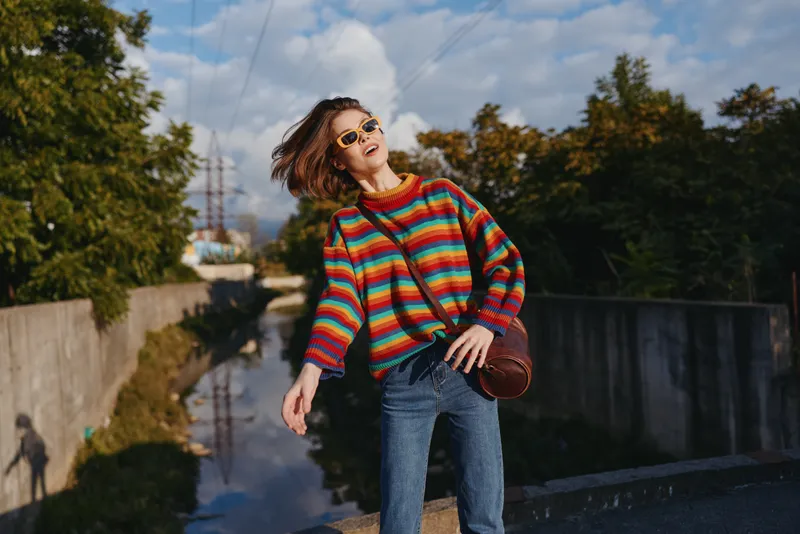 Woman in a colorful striped sweater and sunglasses, smiling with short hair and shoulder bag on an urban canal bridge, leaning back in casual jeans for travel lifestyle portrait.