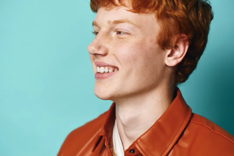 Young man with red hair smiling happily in profile, wearing orange shirt against bright blue background, portrait of joyful expression and casual style.