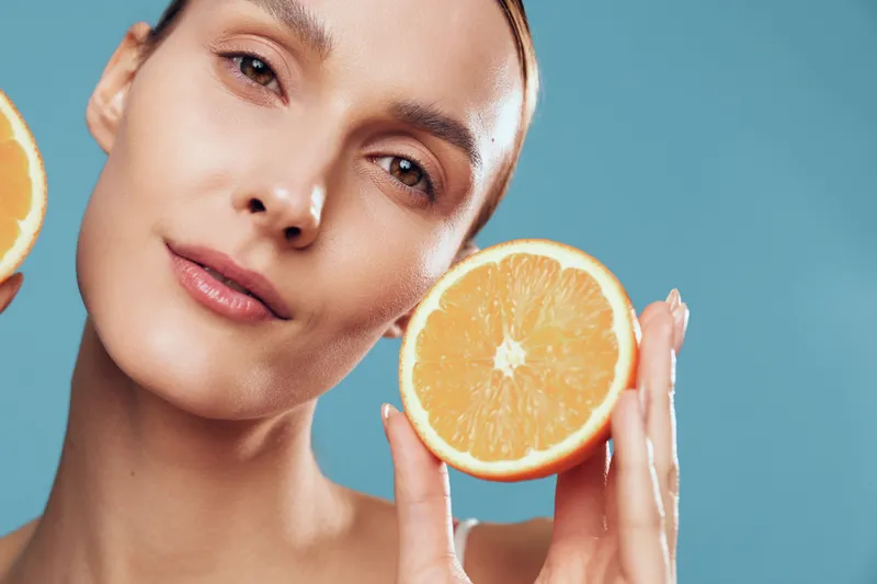 Cheerful woman portrait with glowing skin, holding a fresh orange slice by her cheek, citrus skincare concept, beauty routine, clean blue background, soft natural light, healthy lifestyle