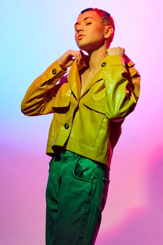 Young woman with short pink hair wearing yellow jacket and green jeans poses confidently against colorful gradient background with warm lighting.