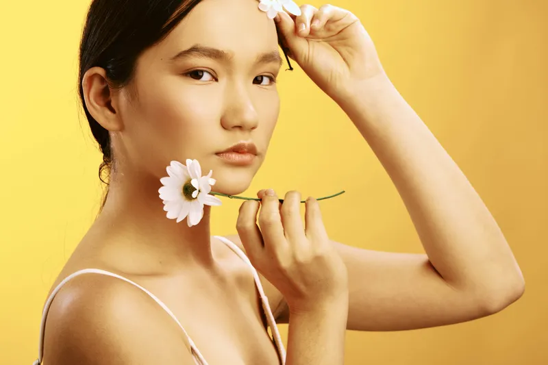 asian woman with delicate features poses gracefully in a vivid studio setting, holding flowers and adjusting a petal, exuding calm elegance and natural beauty.