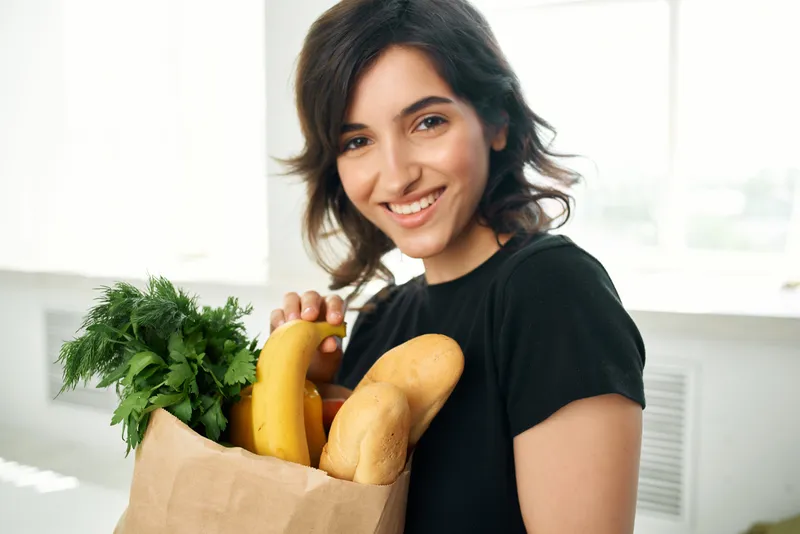 Жизнерадостная женщина с пакетом продуктов делает домашнюю работу, питается здоровой пищей