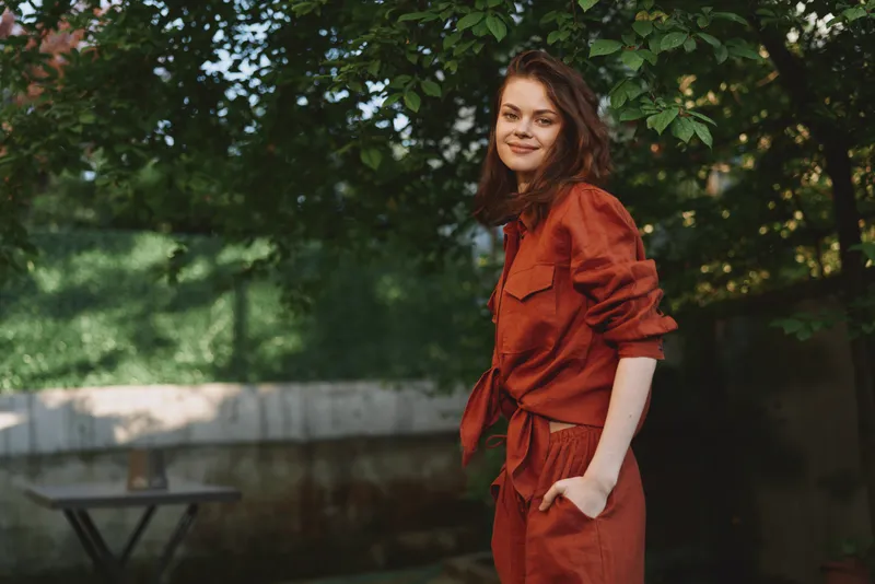 Smiling woman wearing a red outfit stands outdoors in a garden, natural light highlighting style and confidence in a candid lifestyle moment on a clean background
