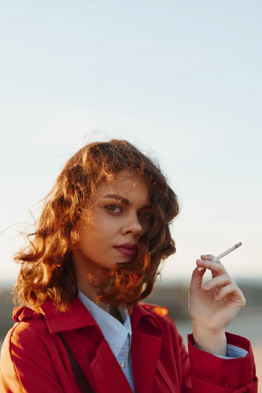A young woman with curly hair wearing a red coat smokes a cigarette outdoors during golden hour with a clear sky background. Concept for lifestyle and contemplation.