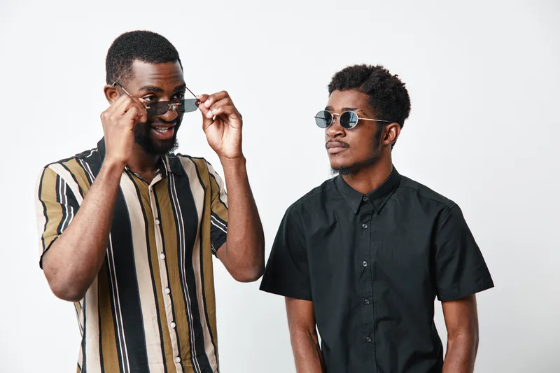 Two young black men with dark skin wearing sunglasses and casual shirts posing together on white background for fashion and friendship concept.