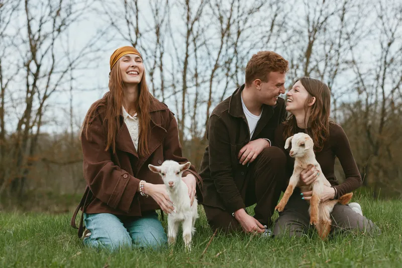 Lifestyle shot of friends outdoors with goats and kids, relaxed atmosphere, smiling trio sits on grass in a park, casual clothing, warm mood, affectionate interaction, animals present, natural light,