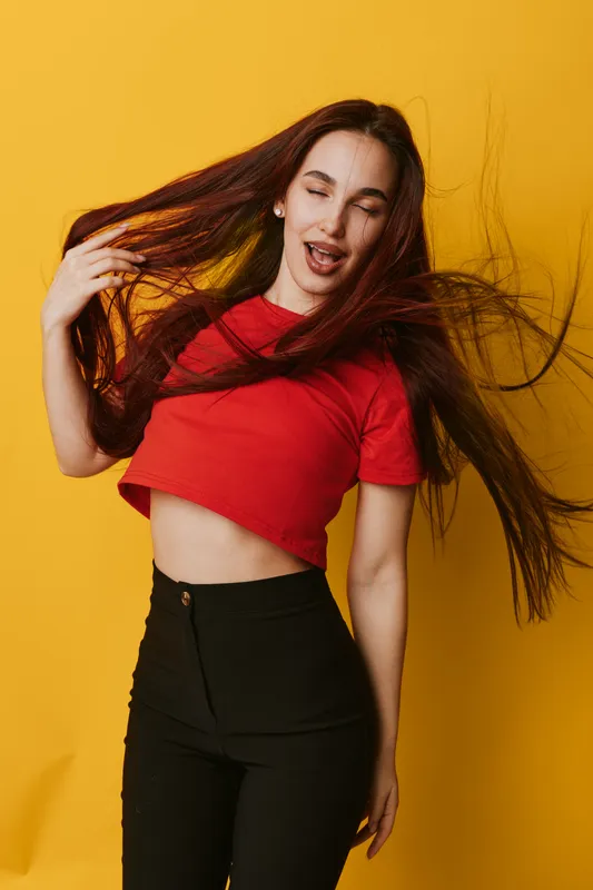 Young woman with long hair wearing red crop top and black pants posing with eyes closed and hair flying on yellow background. Fashion, beauty, casual style and motion concept.