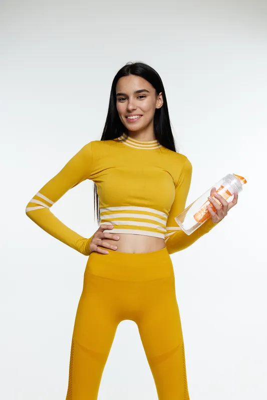 Young woman sport fitness enthusiast in vibrant yellow workout clothes holding a water bottle, smiling confidently with long dark hair against white background.