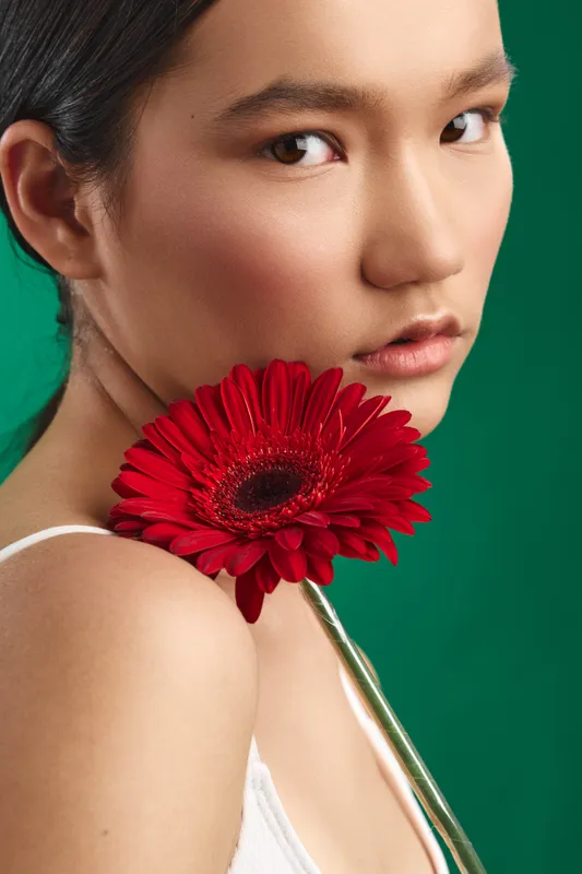 Asian woman appears in a studio portrait, holding a red flower near her shoulder against a vivid green backdrop. The closeup exudes elegance, calm, and refined beauty.