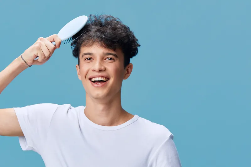 smiling man brushing hair, casual white t-shirt, joyful expression, healthy dark curly hair, blue background, young adult male with carefree mood