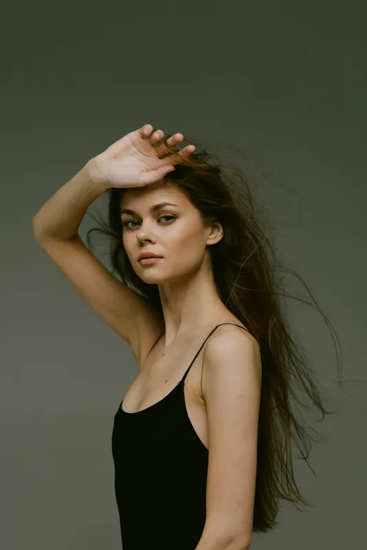 Portrait of a young woman with long hair shielding her eyes, wearing a black tank top against a muted green background.