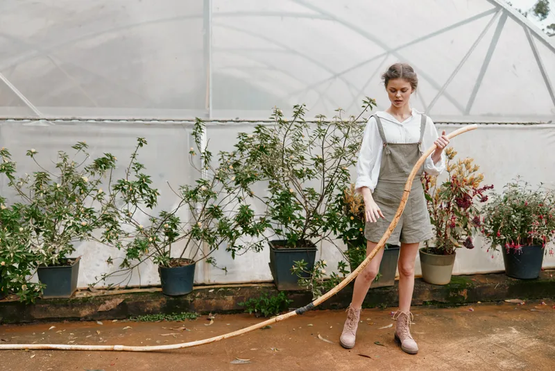Young woman in casual outfit gardening in greenhouse, showcasing a serene expression and natural beauty among lush plants, emphasizing seasonal planting and sustainability