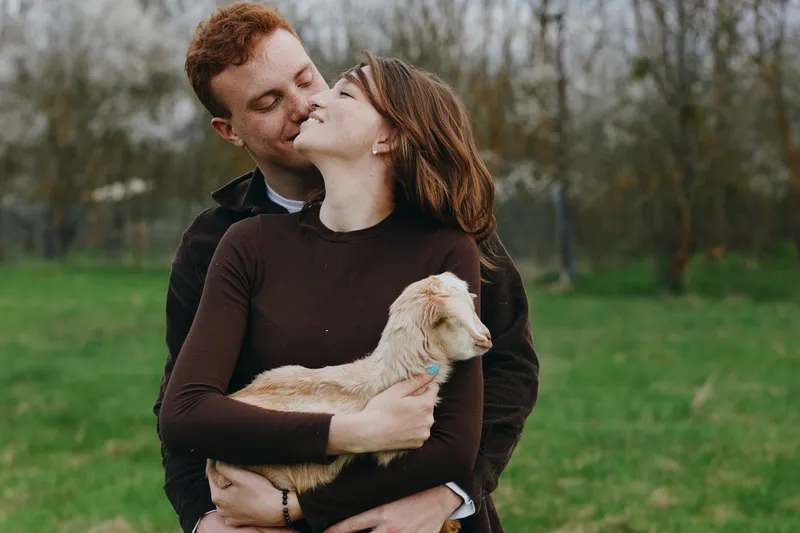 A couple outdoors share a tender kiss while cradling a small goat kid, creating a warm, candid scene of love and animal companionship in a sunny field.