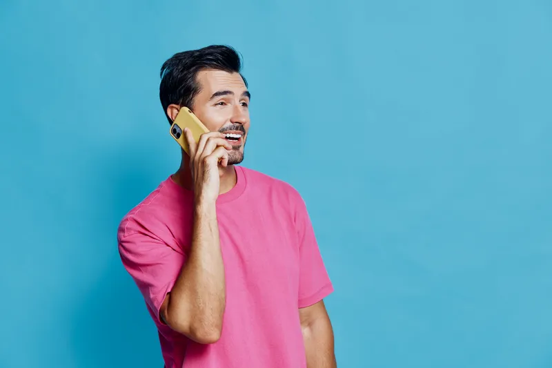Smiling man wearing pink t-shirt talking on mobile phone against a bright blue background. Cheerful and vibrant lifestyle concept for communication and connection.