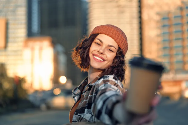 Woman smile coffee urban beanie golden hour glow, candid lifestyle portrait of a happy young woman offering a takeaway cup on a city street, authenticity and mindful living in warm light.