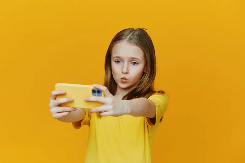 A young girl with brown hair takes a selfie against a vibrant yellow background, exuding a playful expression that signifies joy and enthusiasm