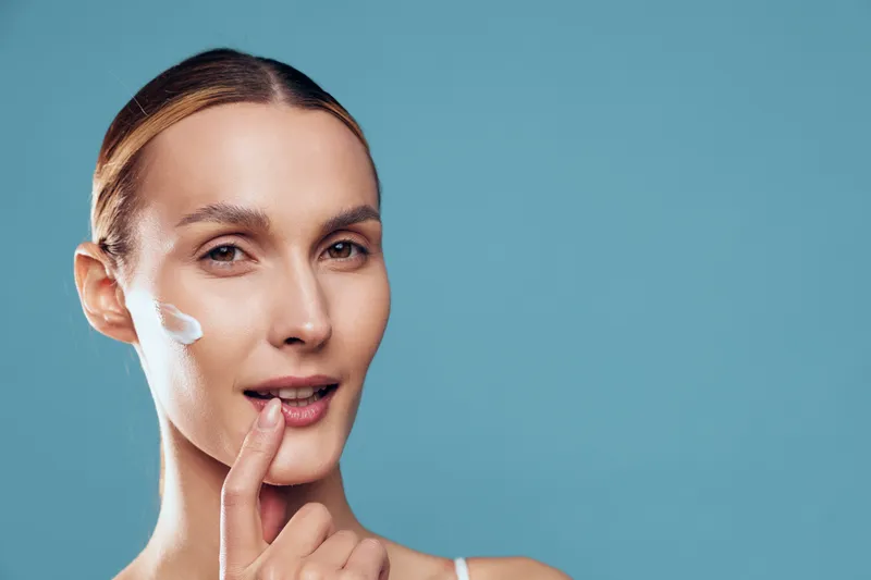Young woman with clear skin applying white face cream on cheek, looking at camera with soft smile, blue background, concept of skincare, beauty, wellness, self-care, and health.