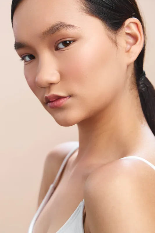 Asian woman gazes softly at the camera in a clean studio setting, exuding calm confidence, graceful poise, and timeless beauty through a simple, elegant pose.