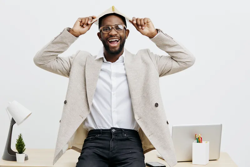joyful professional man wearing glasses and beige blazer holds a white paper hat above his head while smiling, sitting at a modern office desk with laptop, stationery, and a small potted plant, indoor