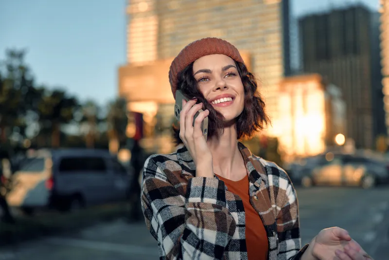 Woman with phone and smile in an urban street, candid authenticity and golden hour lifestyle glow creating mindful living vibe; emotional storytelling portrait of modern city life.
