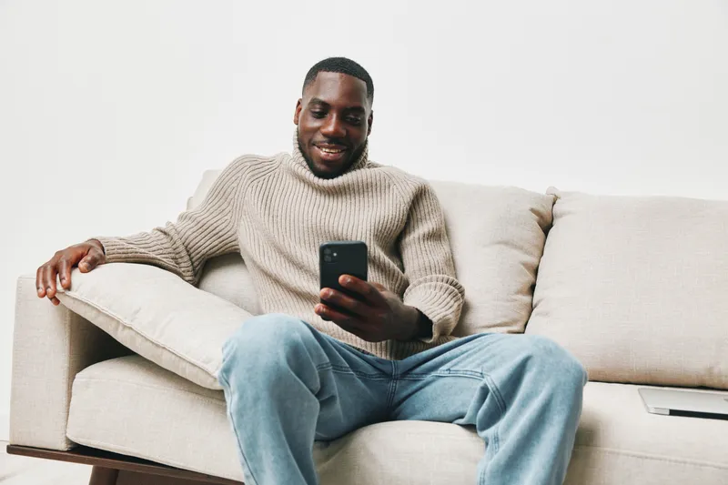 Smiling Black man in cozy sweater using smartphone on sofa in a modern living room, showcasing relaxation and connection in a contemporary space