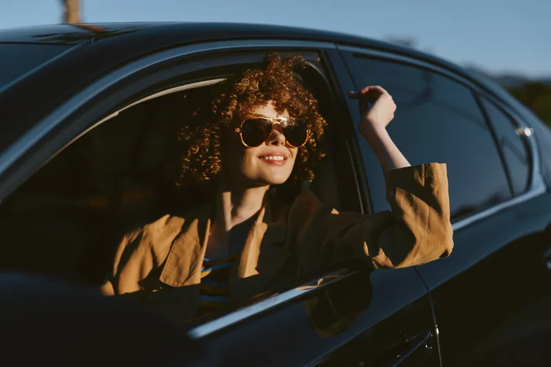 Young woman in a car holds the window frame, wearing sunglasses and retro clothing, candid lifestyle image with warm film tones, stylish everyday moment captured.