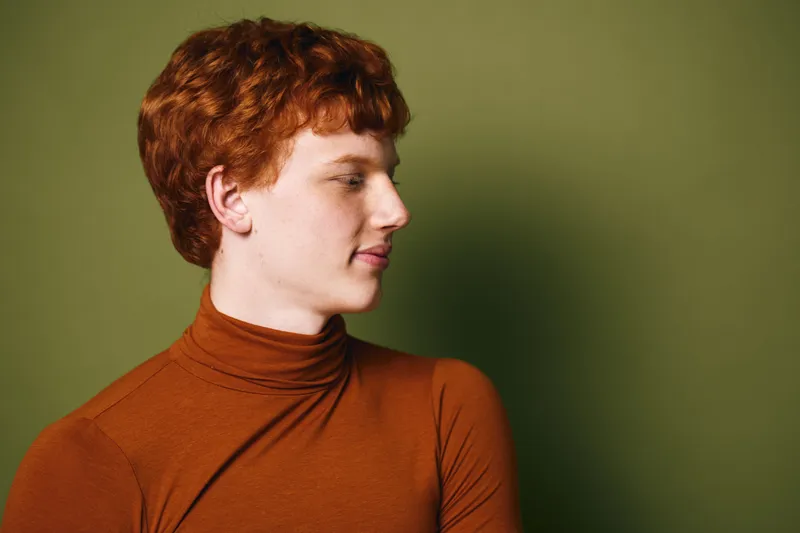 Young man with red hair and fair skin in orange turtleneck looking sideways against green background. Portrait, profile, calm expression and casual style with studio lighting and minimalism.