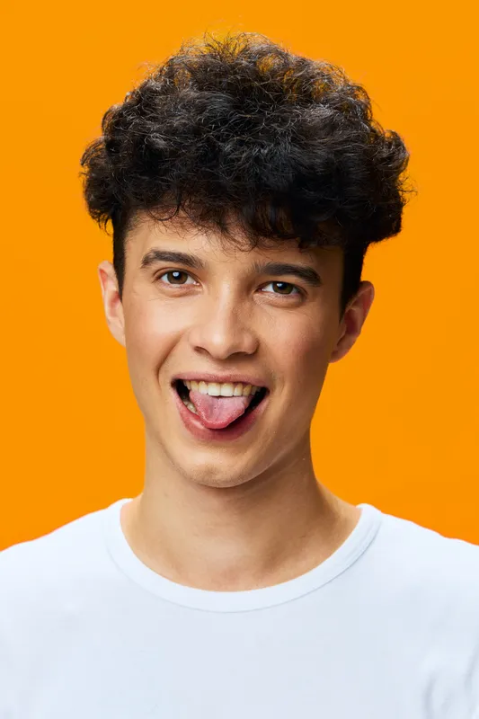 young man sticking out tongue wearing white t-shirt on bright orange background showing playful and fun attitude with curly hair