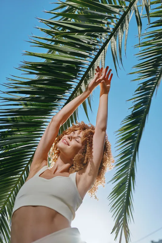 smiling woman with curly hair stretches arms under tropical palm leaves in bright sunlight and blue sky, wearing white top during summer day outdoors.
