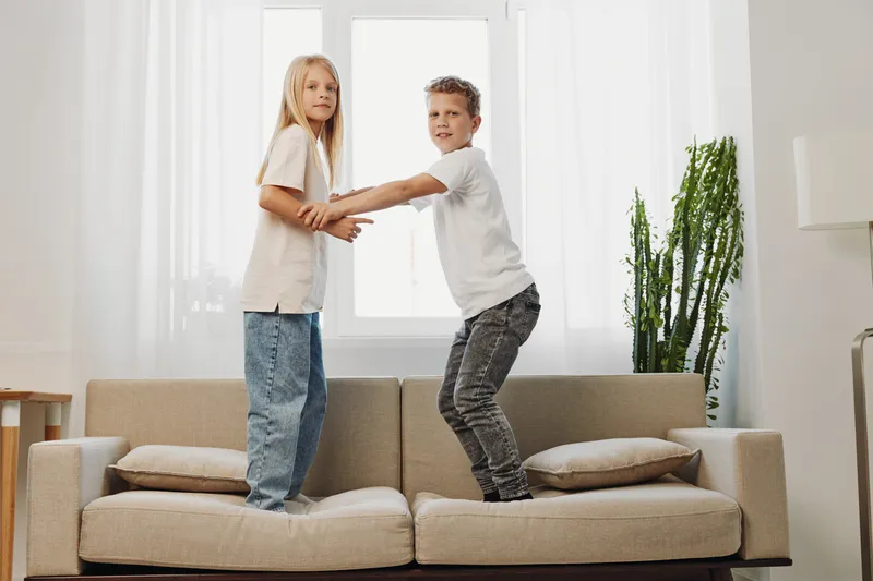Two children standing on a beige sofa holding hands in a bright living room with natural light. Concept of playful siblings, family bonding, and childhood joy indoors.