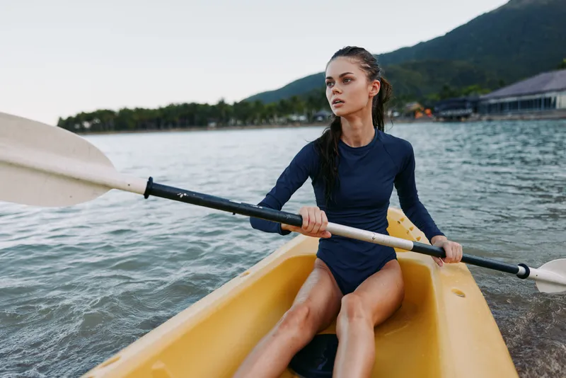A focused female kayaker paddles a yellow boat along a calm coastal inlet, wearing a navy long sleeve swimsuit, with braided hair and distant hills.