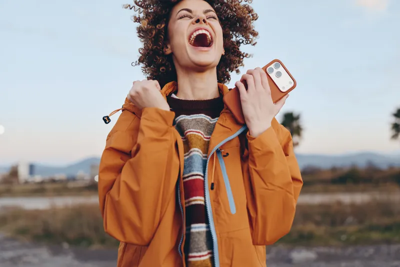 Woman lifestyle outdoors laughing, holding smartphone, orange jacket and scarf, curly hair lifted by breeze, autumn sunlight, joyful expression, blurred road and landscape background