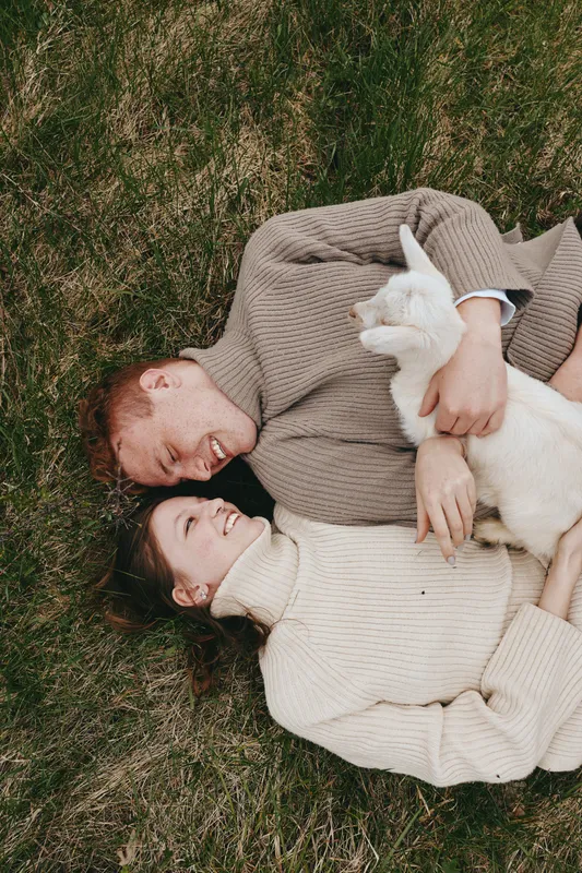 Couple lounging on grass in cozy sweaters, a gentle goat kid resting beside them, showing affection, laughter, and warm outdoor vibes in a natural meadow setting and shared warmth together