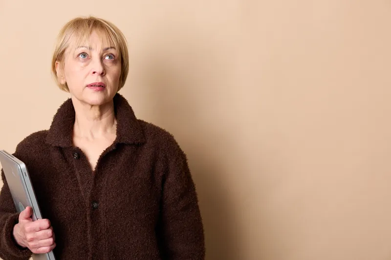 Senior woman holding smartphone, surprised worried face, looking at screen, indoor portrait on beige wall, dark coat, soft light, copyspace for text, modern mobile communication