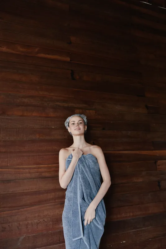 woman wrapped in towel standing against wooden wall with towel turban on head in spa setting, calm expression, warm lighting