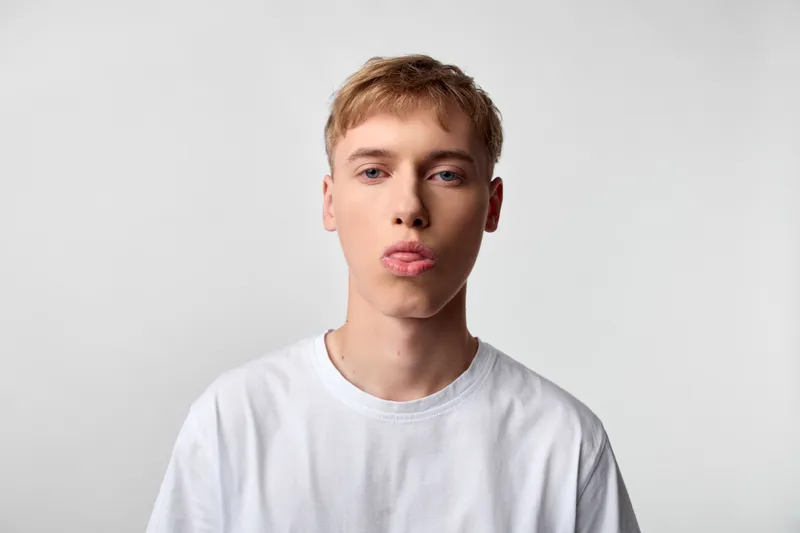 Tongue out, young man in white t shirt, playful expression in a studio setting. Close crop highlights casual mood, soft lighting and minimal background for versatile lifestyle, fashion or teen