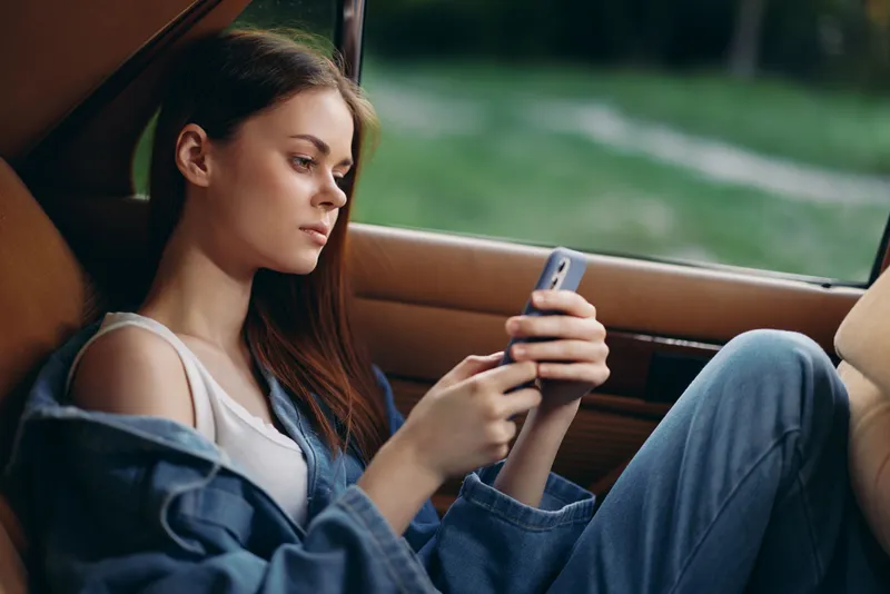 Young woman in casual denim attire relaxing in the car, using her smartphone, showcasing a serene expression against a natural background.