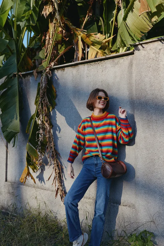 Woman in mid twenties wearing a colorful striped sweater and sunglasses, smiling while leaning against a concrete wall with banana leaves, casual outfit, travel lifestyle, relaxed city tropics.