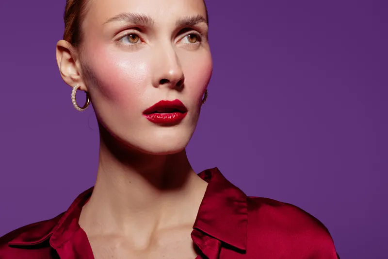 Close-up portrait of a woman with red lipstick and hoop earrings wearing a red blouse on purple background. Concept of beauty, fashion, and makeup with dramatic lighting.