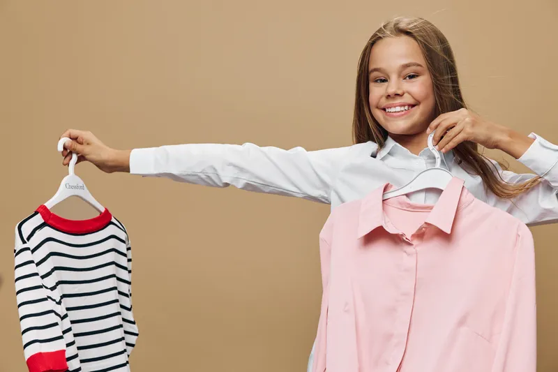 Smiling girl holding two clothes hangers with striped t shirt and pink blouse standing on beige background. Teenager with long hair choosing outfit for casual or formal wear.