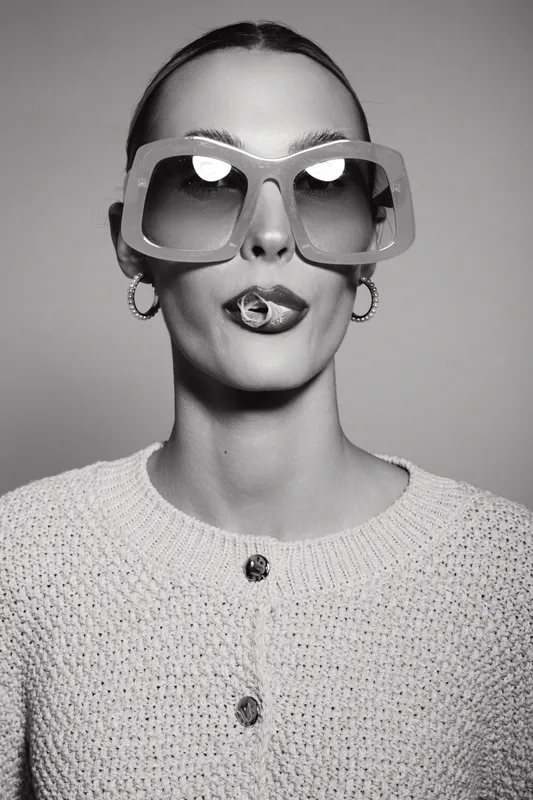 Portrait of white woman in oversized sunglasses with candy on tongue wearing knitted sweater. Minimal studio background.