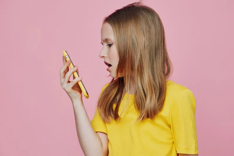 Surprised girl in a yellow t shirt looking at her phone against a pink background, expressing curiosity and excitement, perfect for social media themes